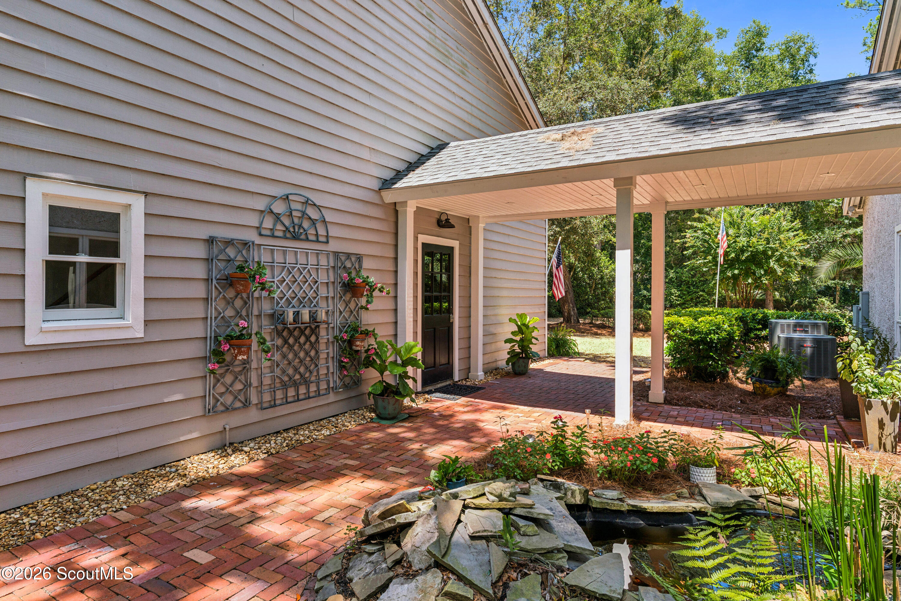 Covered brick patio beside the home, with trellis planters and a small rock-edged pond.