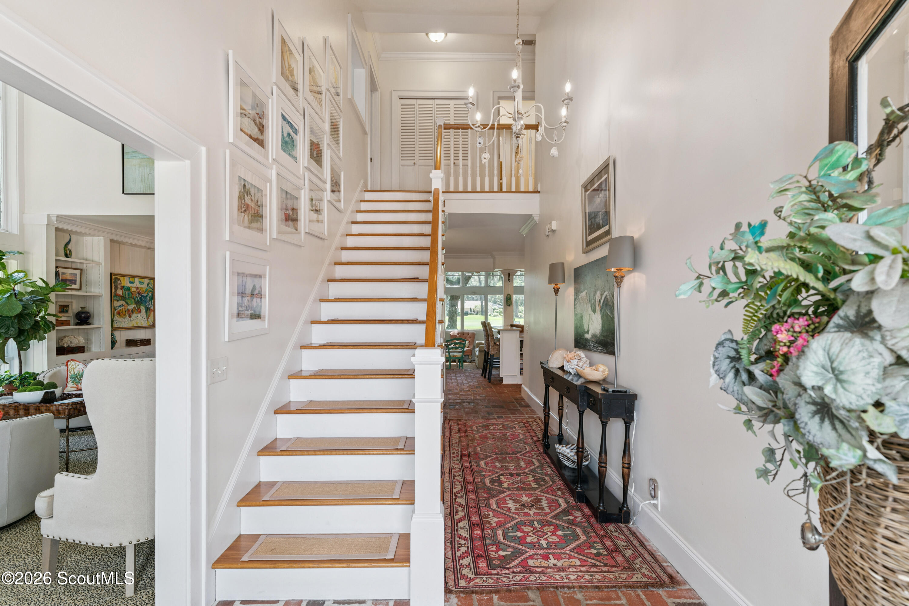 Bright foyer with a white staircase, framed art, chandelier, and patterned runner over brick flooring.