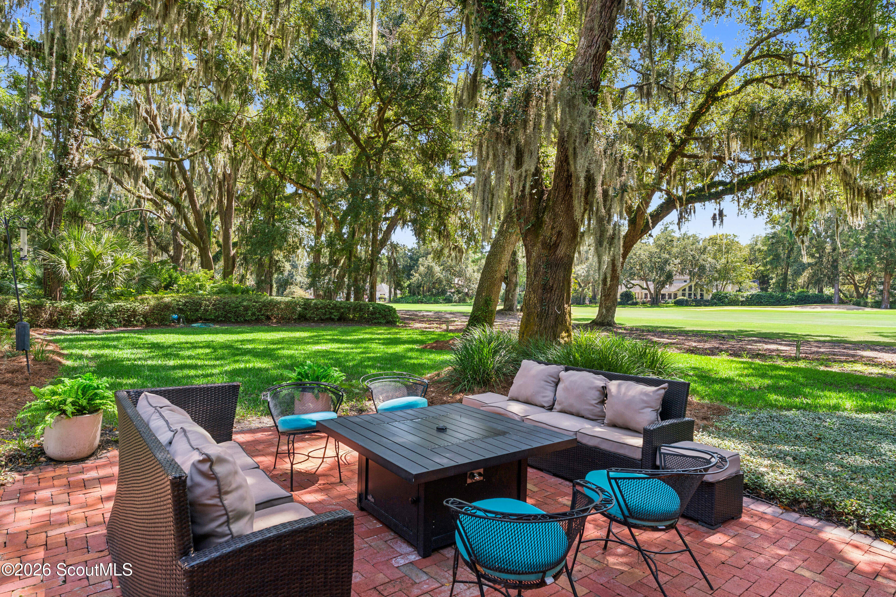 Brick patio with wicker seating beneath moss-draped oaks beside a broad green lawn.