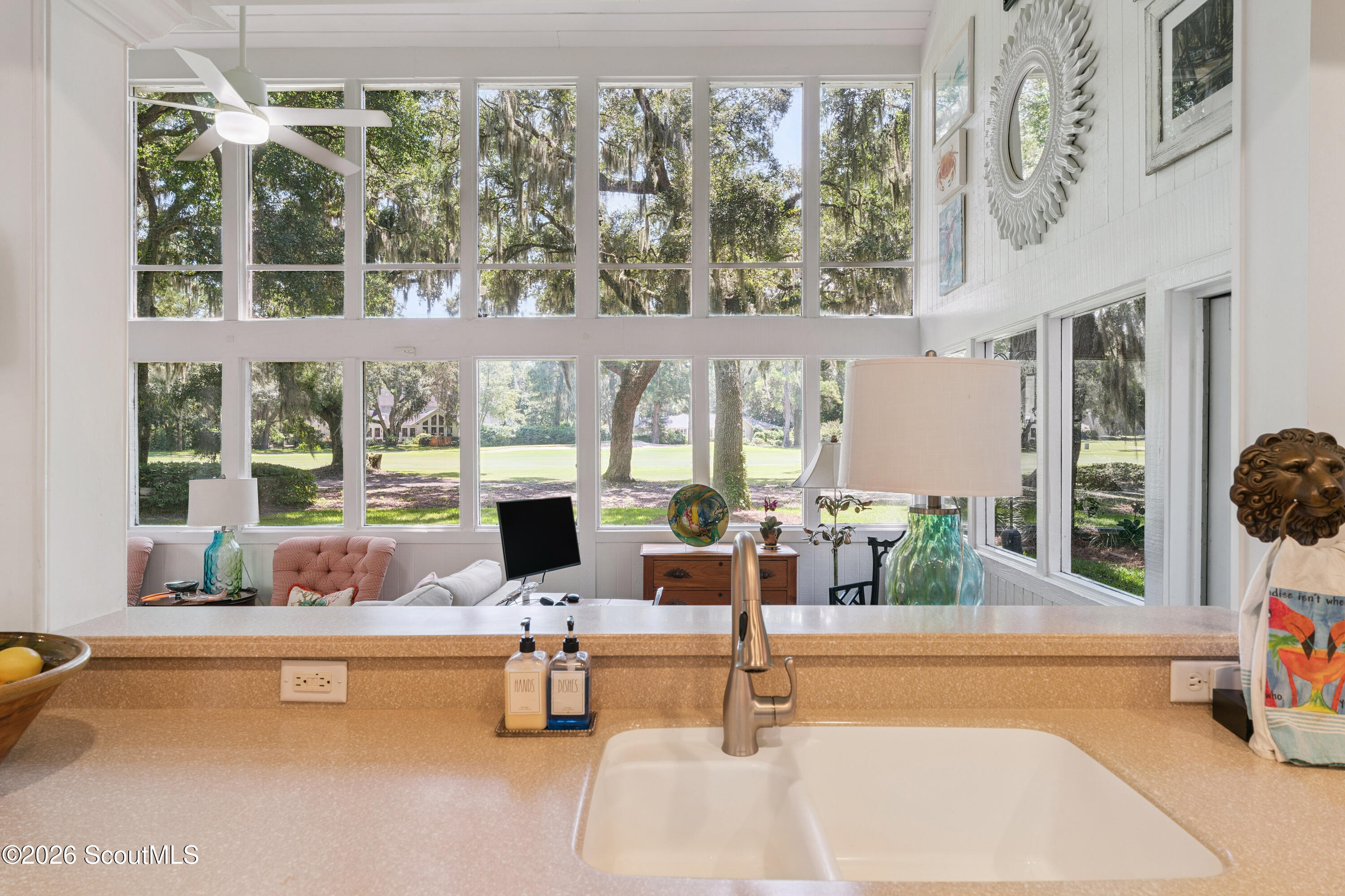 Bright kitchen sink and counter overlooking a window-filled sitting area and leafy lawn.