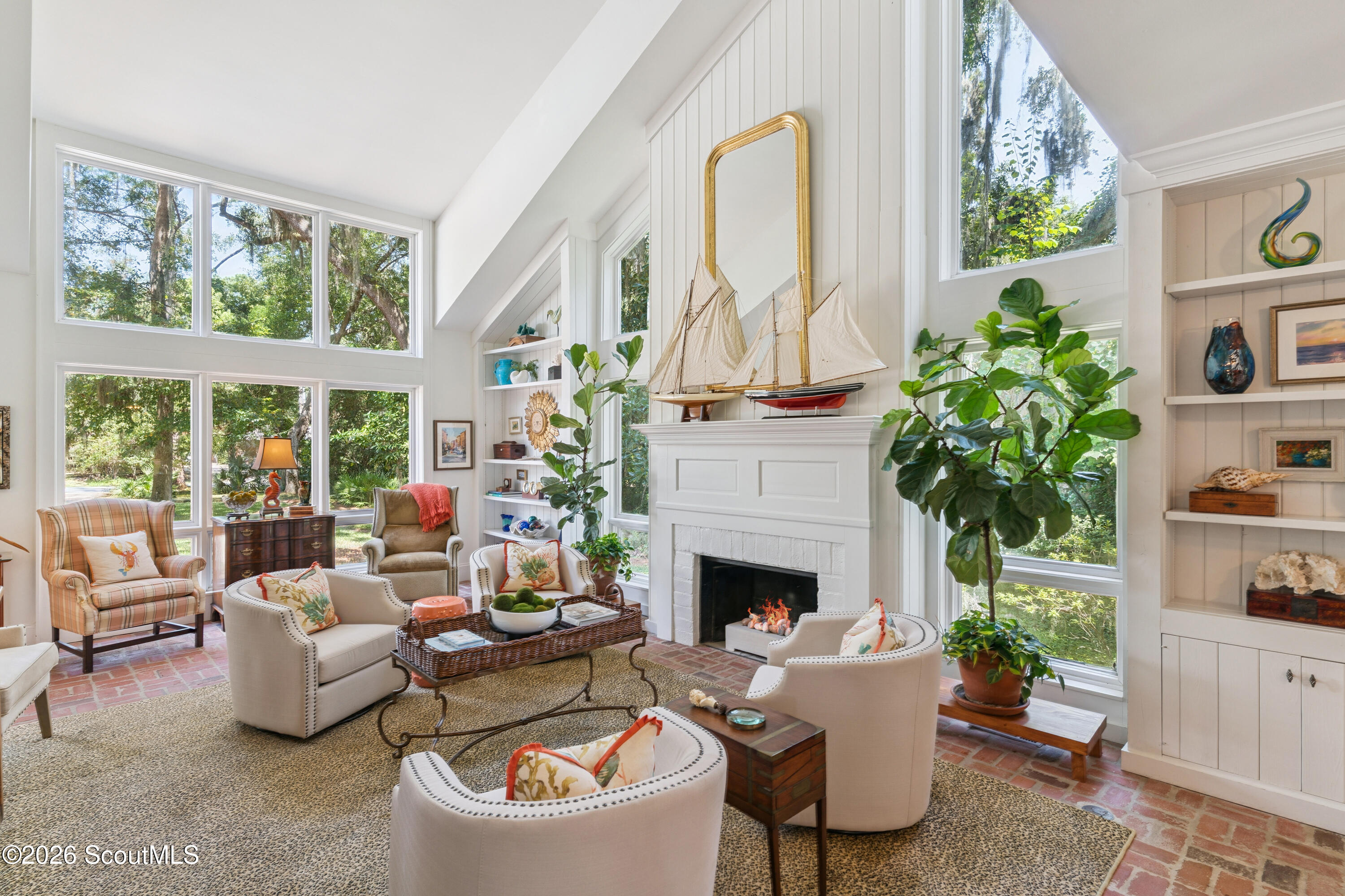 Bright living room with a white fireplace, tall tree-view windows, built-in shelves, and sailboat models.