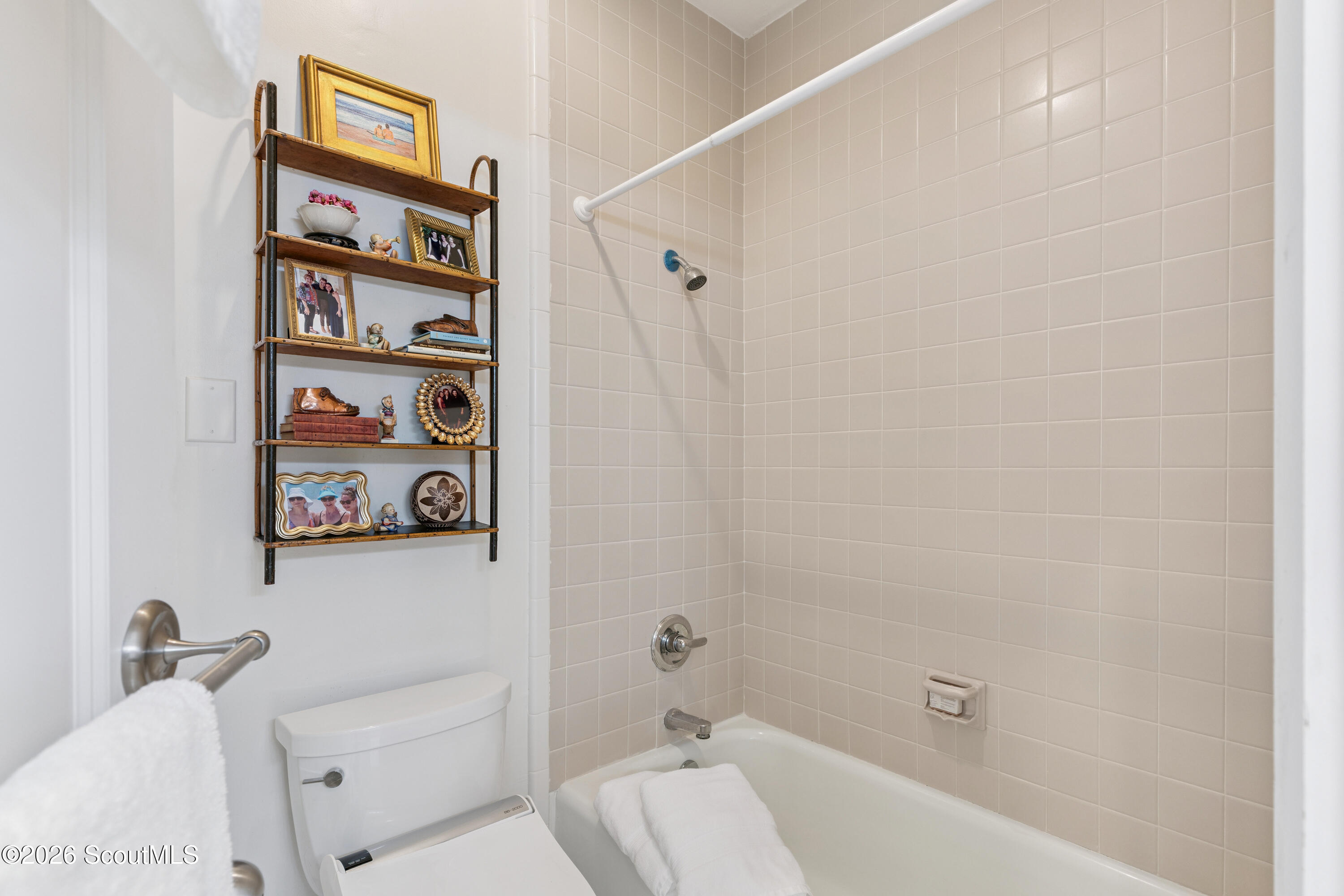 Bathroom with beige tiled tub and shower beside a toilet and photo-filled wall shelf.