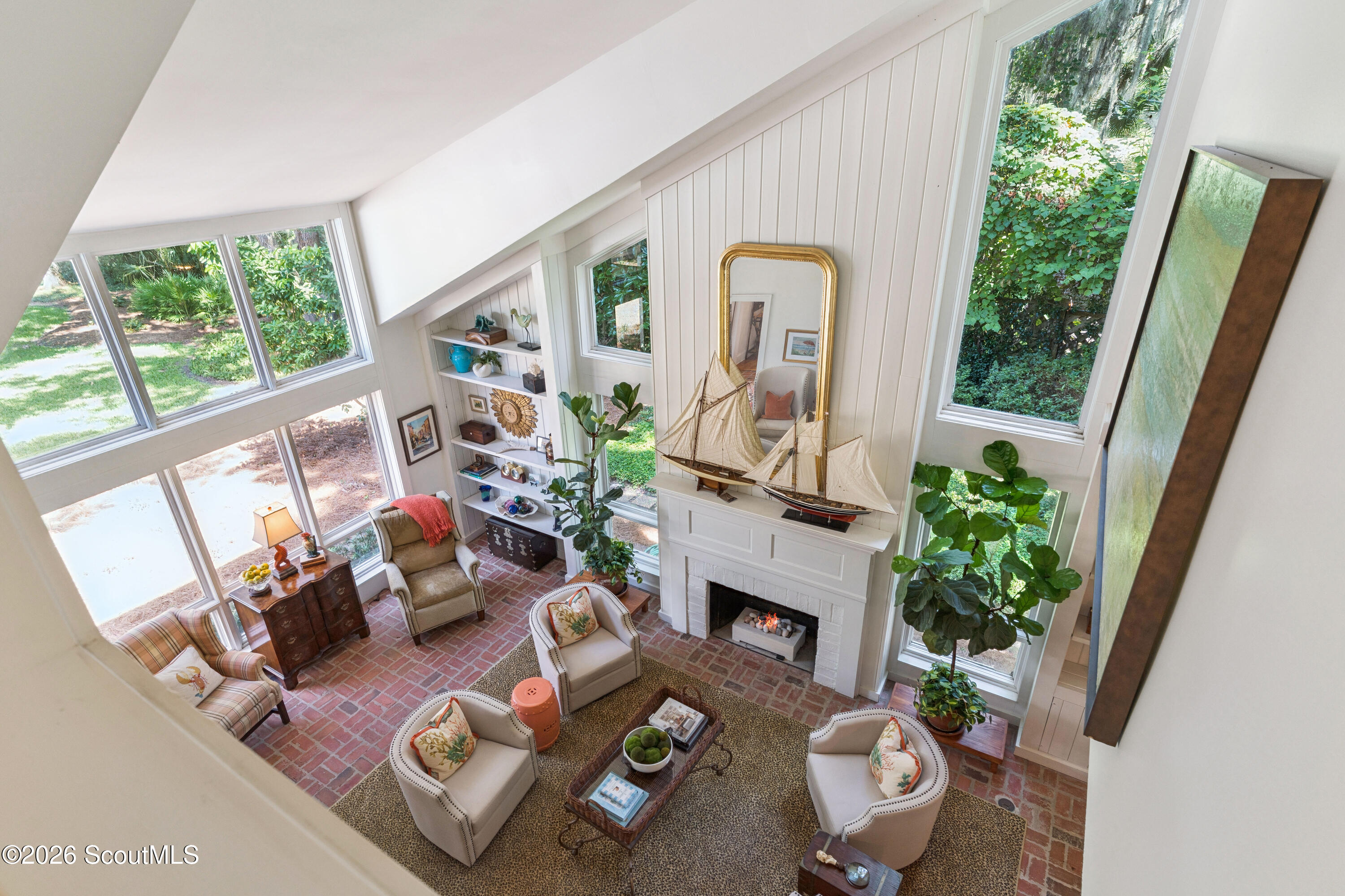 Living room with a high sloped ceiling, tall windows, brick floor, and white fireplace topped with model sailing ships.