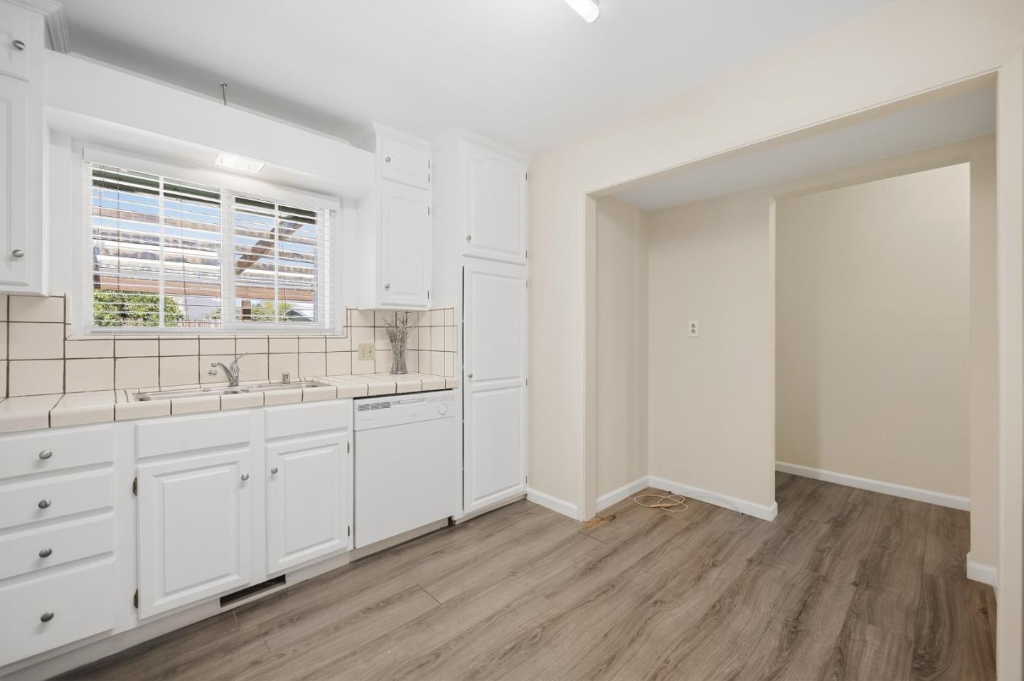 Empty kitchen with white cabinets, tiled counters, a window above the sink, and plank flooring.
