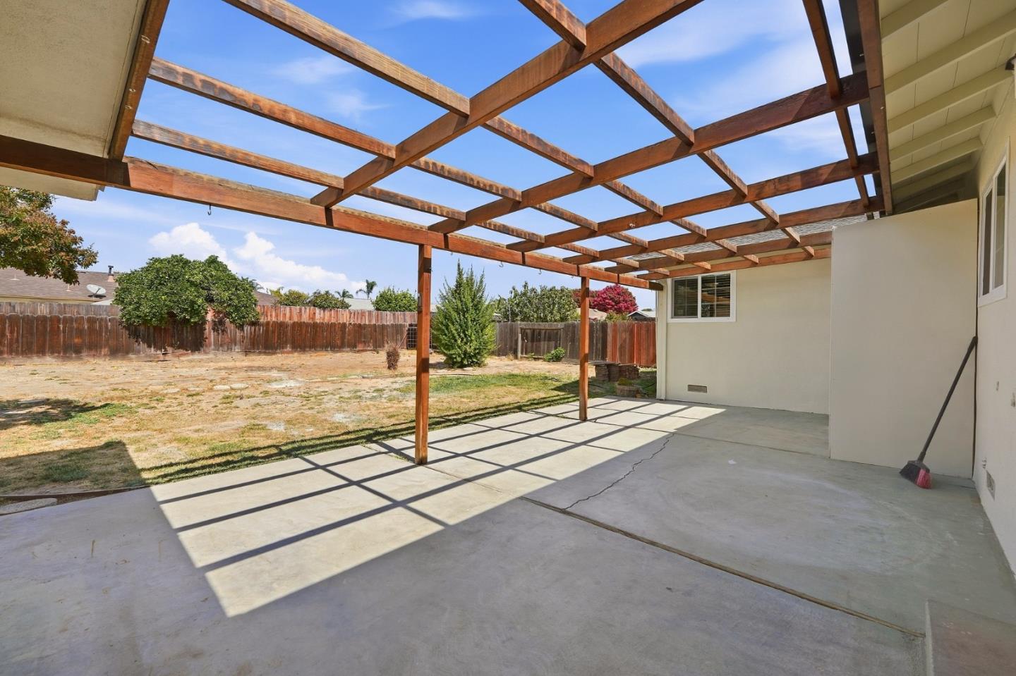 Concrete patio beneath a wooden pergola, overlooking a fenced backyard.
