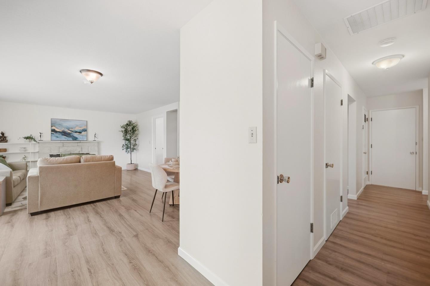 Staged living room and dining area beside a hallway, with beige sofas and light wood-look floors.
