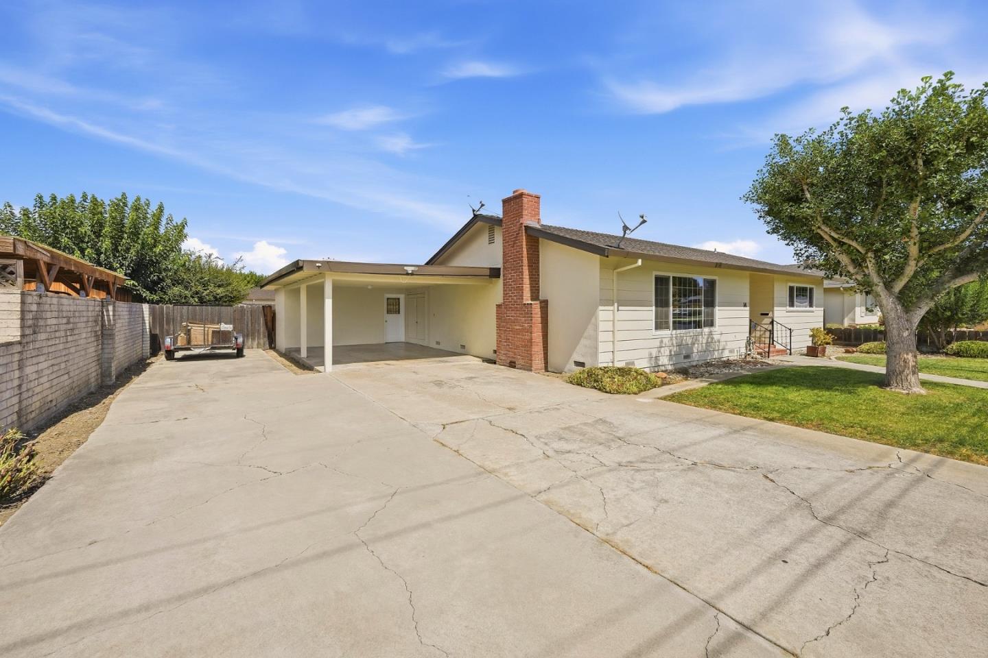 Single-story home with a brick chimney, covered carport, wide driveway, and mature tree.