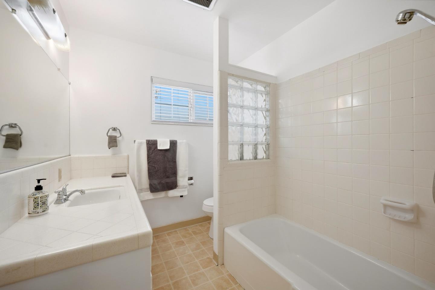 Bathroom with a tiled vanity, bathtub, glass-block window, and hanging towels.