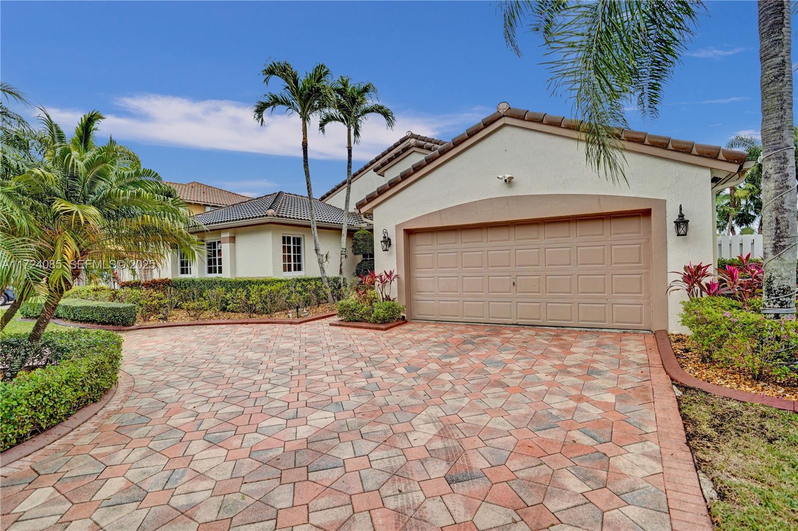 Single-story home with a wide garage, patterned paver driveway, and tropical landscaping.