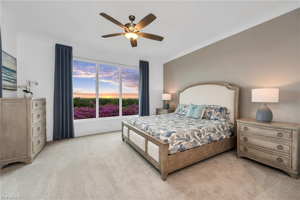 Bedroom with an upholstered bed and wide window overlooking flowering shrubs at sunset.
