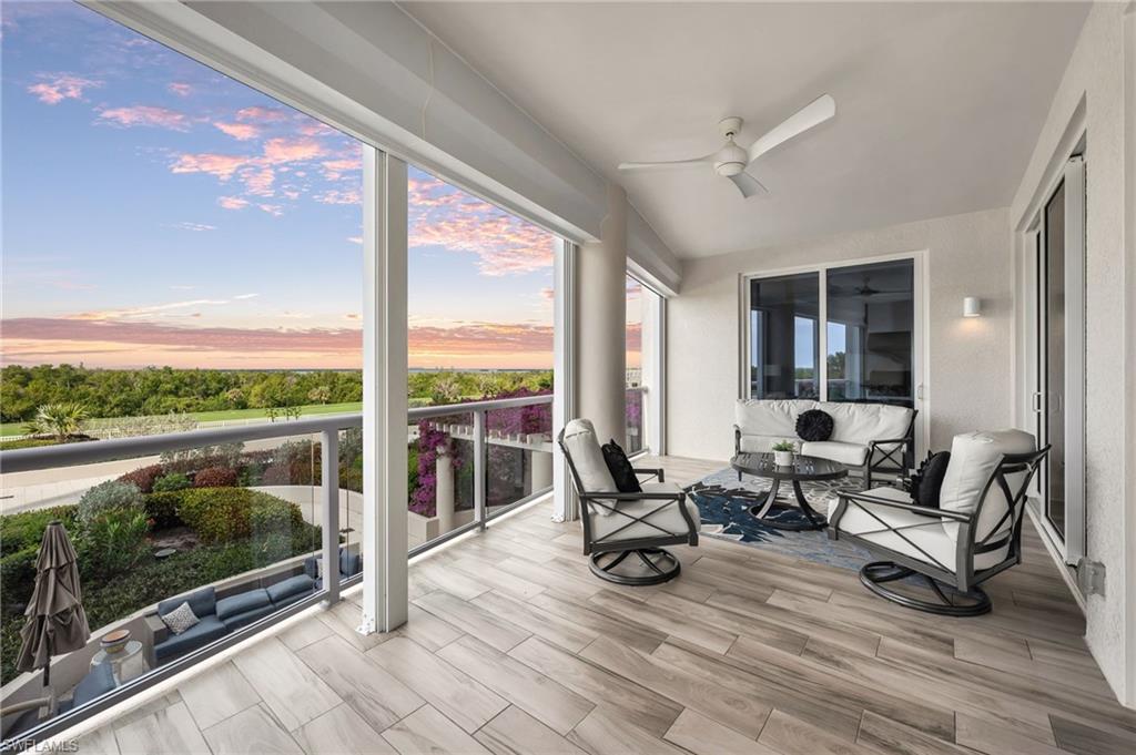 Furnished condo balcony with glass railing, tiled floor, and sunset view over landscaped grounds.