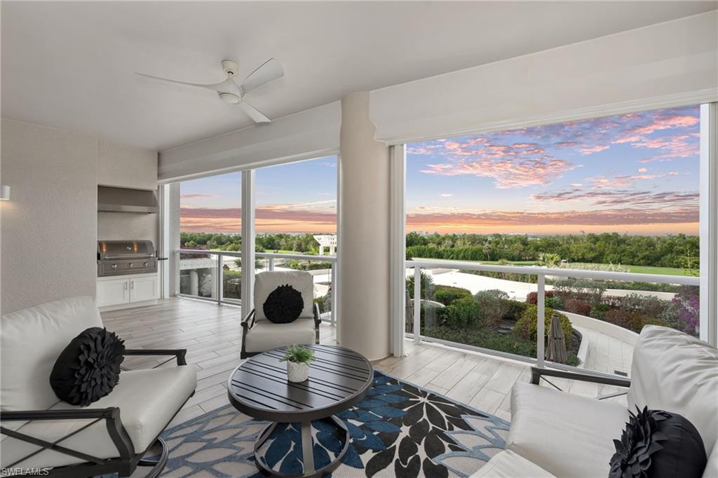 Living room with white seating and floor-to-ceiling windows overlooking a balcony and sunset-lit greenery.