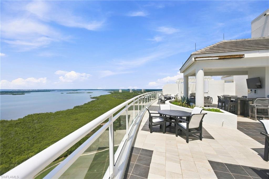 Furnished condo terrace overlooking a broad bay, with glass railing and outdoor dining tables.