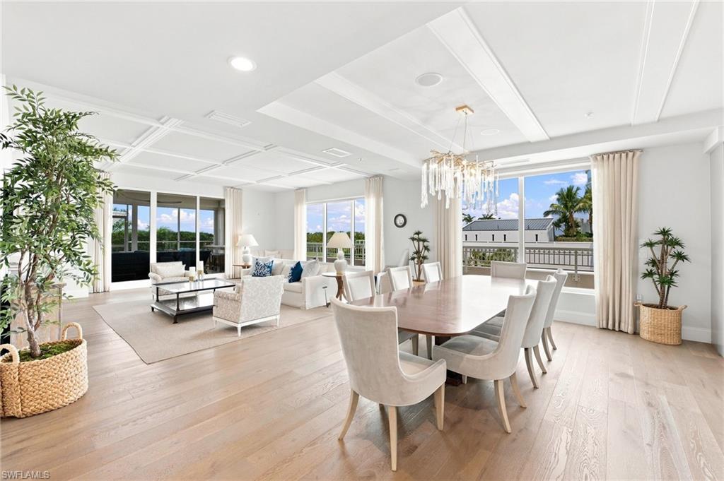 Open living and dining room with a chandelier, pale floors, and broad windows.