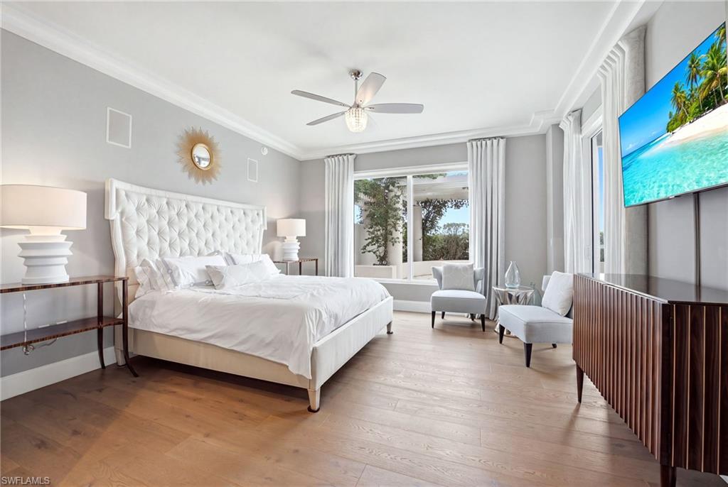 Staged bedroom with a white tufted bed, light wood floors, and seating beside large windows.