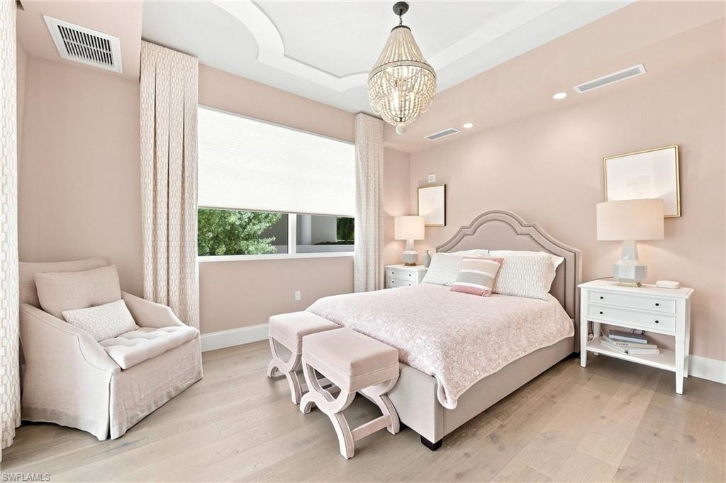Furnished bedroom with pale pink bedding, a crystal chandelier, and an upholstered armchair beside the window.