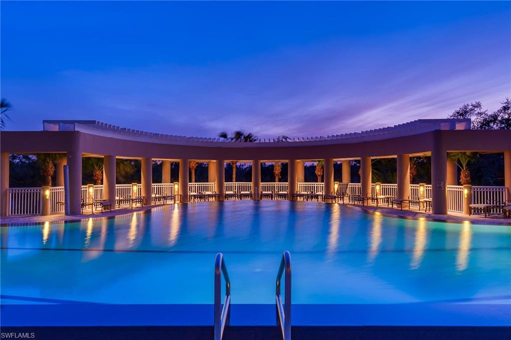 Swimming pool at dusk beneath a curved, columned walkway with warm lights reflected on the water.