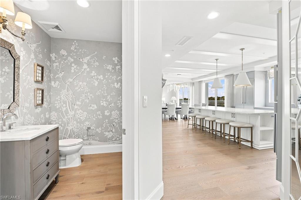 Bright condo kitchen with a long island and bar stools, beside a bathroom with floral wallpaper.