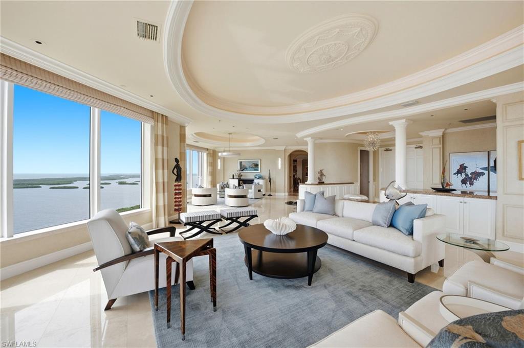 Living room with white furnishings, ornate ceilings, and expansive windows overlooking the water.