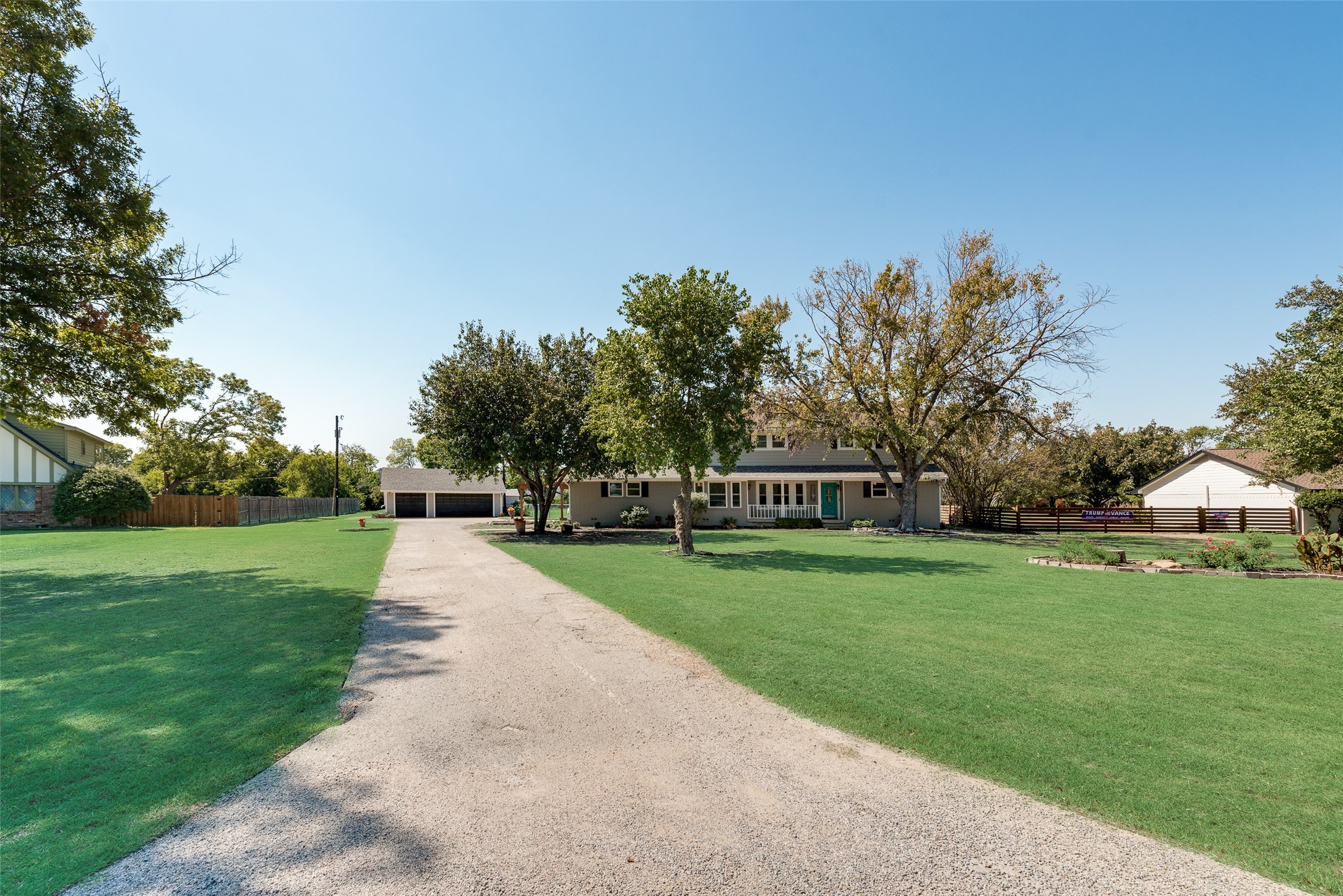 Two-story house with a turquoise door, gravel driveway, detached garage, and broad tree-shaded lawn.