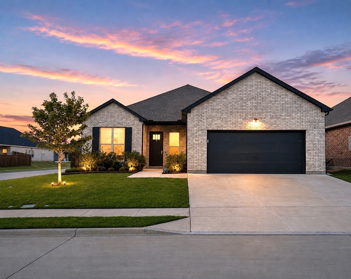 Light brick home with a dark two-car garage and illuminated landscaping beneath a pink dusk sky.