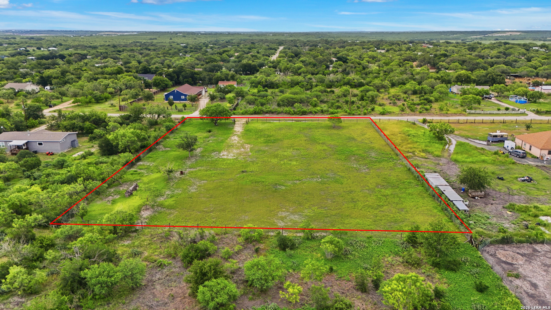 Aerial view of a grassy vacant lot outlined in red, with neighboring homes and trees.