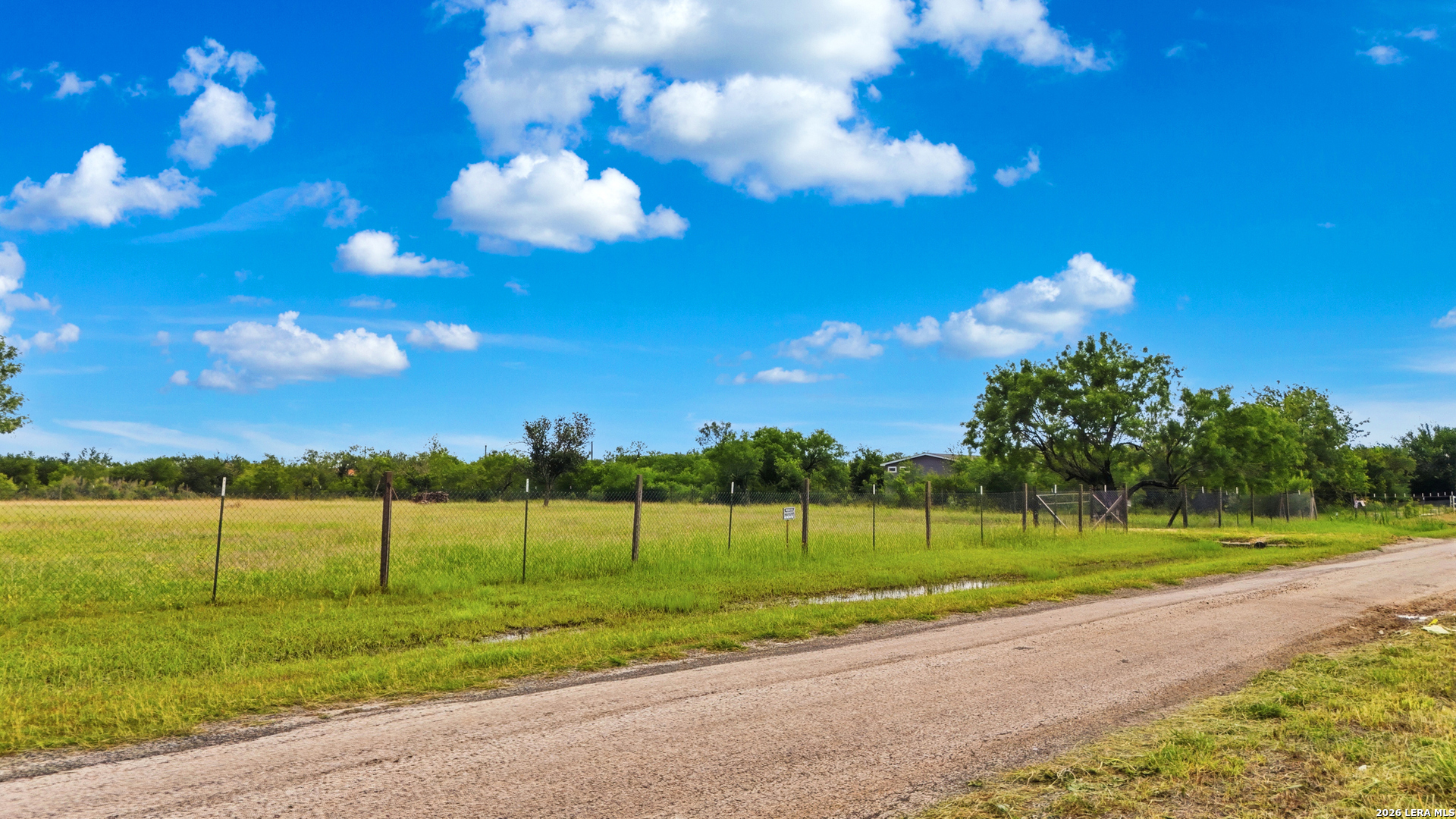 Fenced grassy property beside a rural gravel road, with mature trees and a small building in the distance.