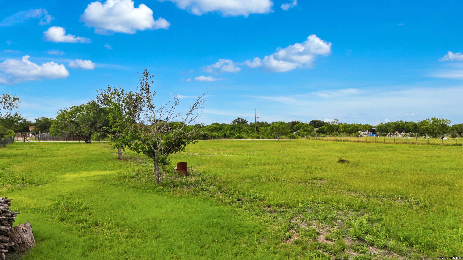 Open grassy property with scattered trees and fenced areas beneath a blue, cloudy sky.