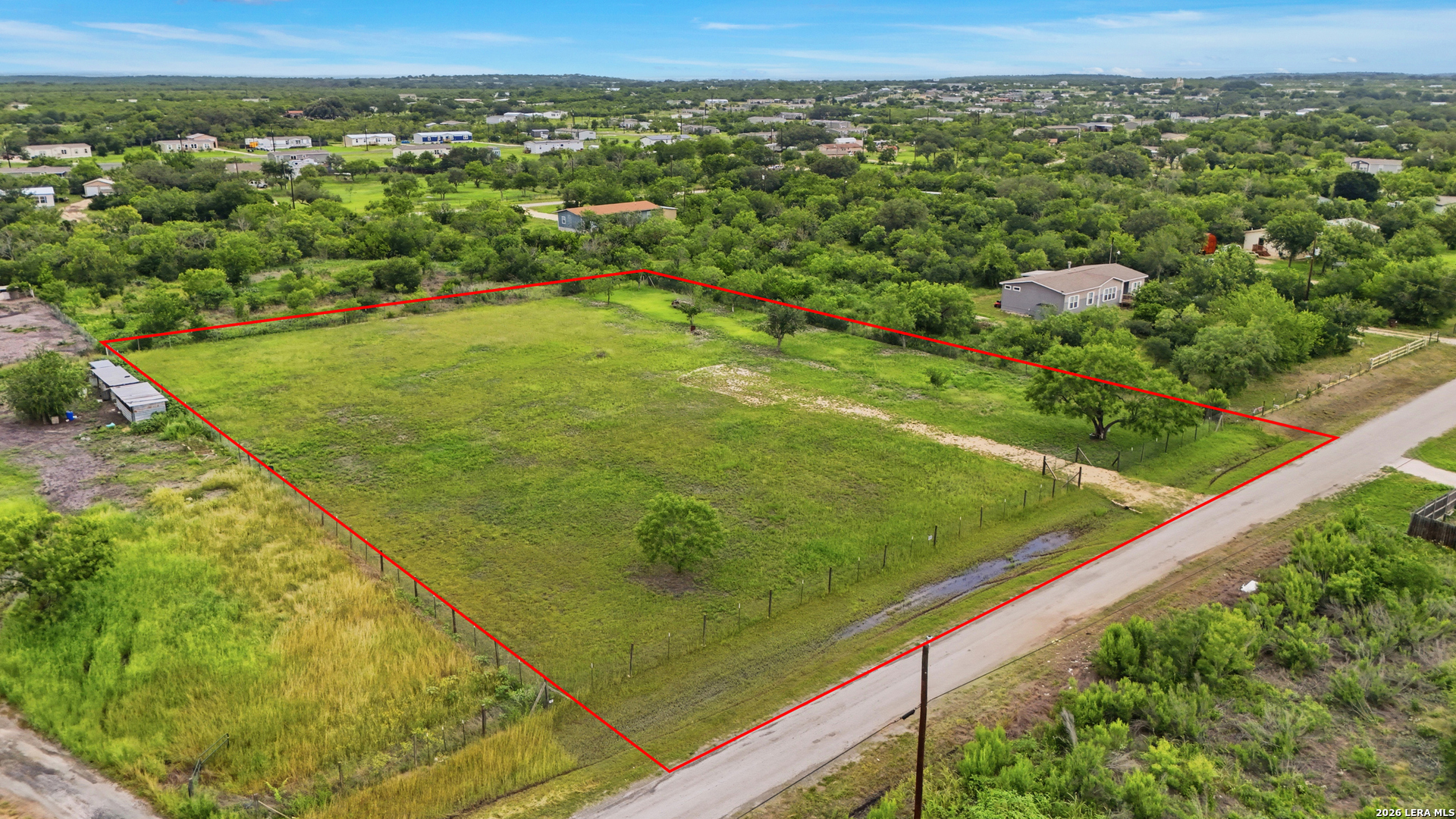Aerial view of a fenced grassy lot outlined in red beside a road.
