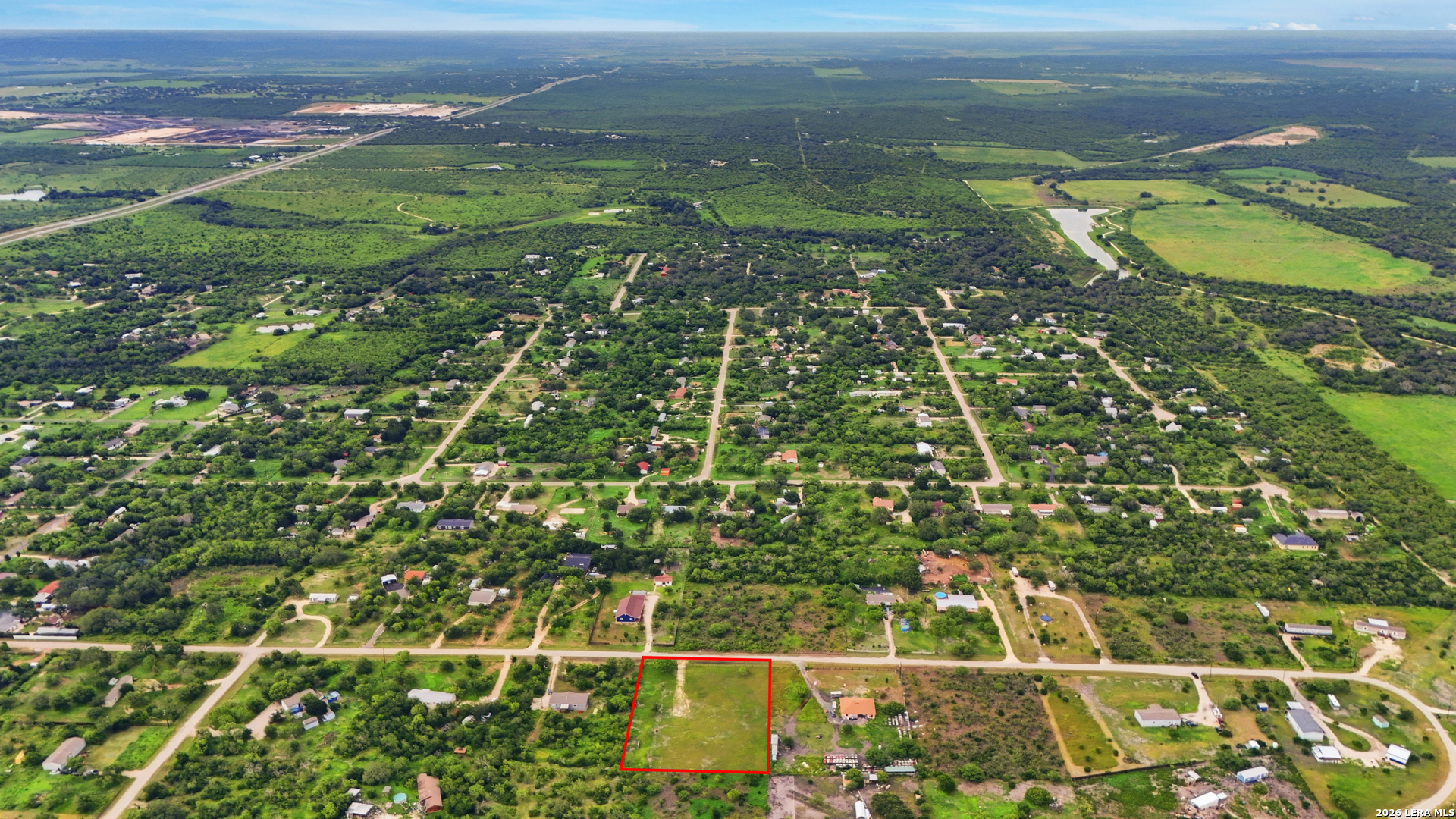 Aerial view of an open grassy parcel outlined in red among rural homes and trees.