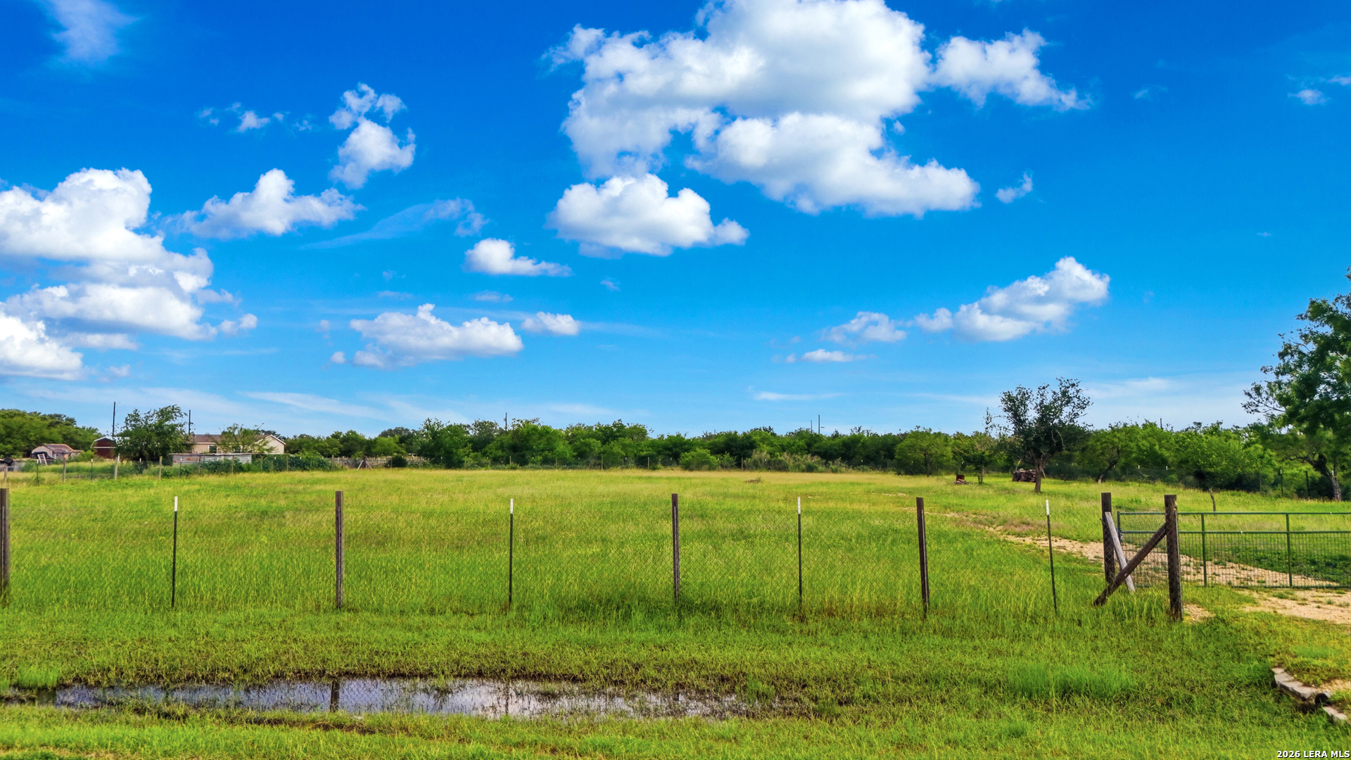 Fenced grassy rural land with a gate and shallow water-filled ditch in the foreground.