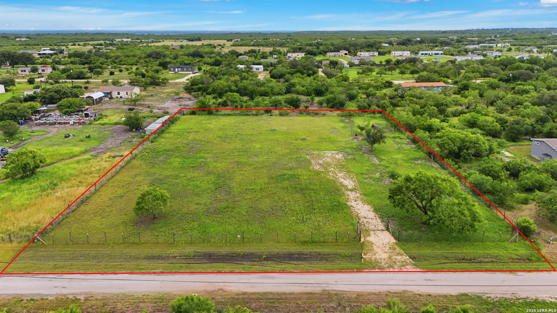 Aerial view of a grassy vacant lot outlined in red, with a dirt driveway and surrounding homes.