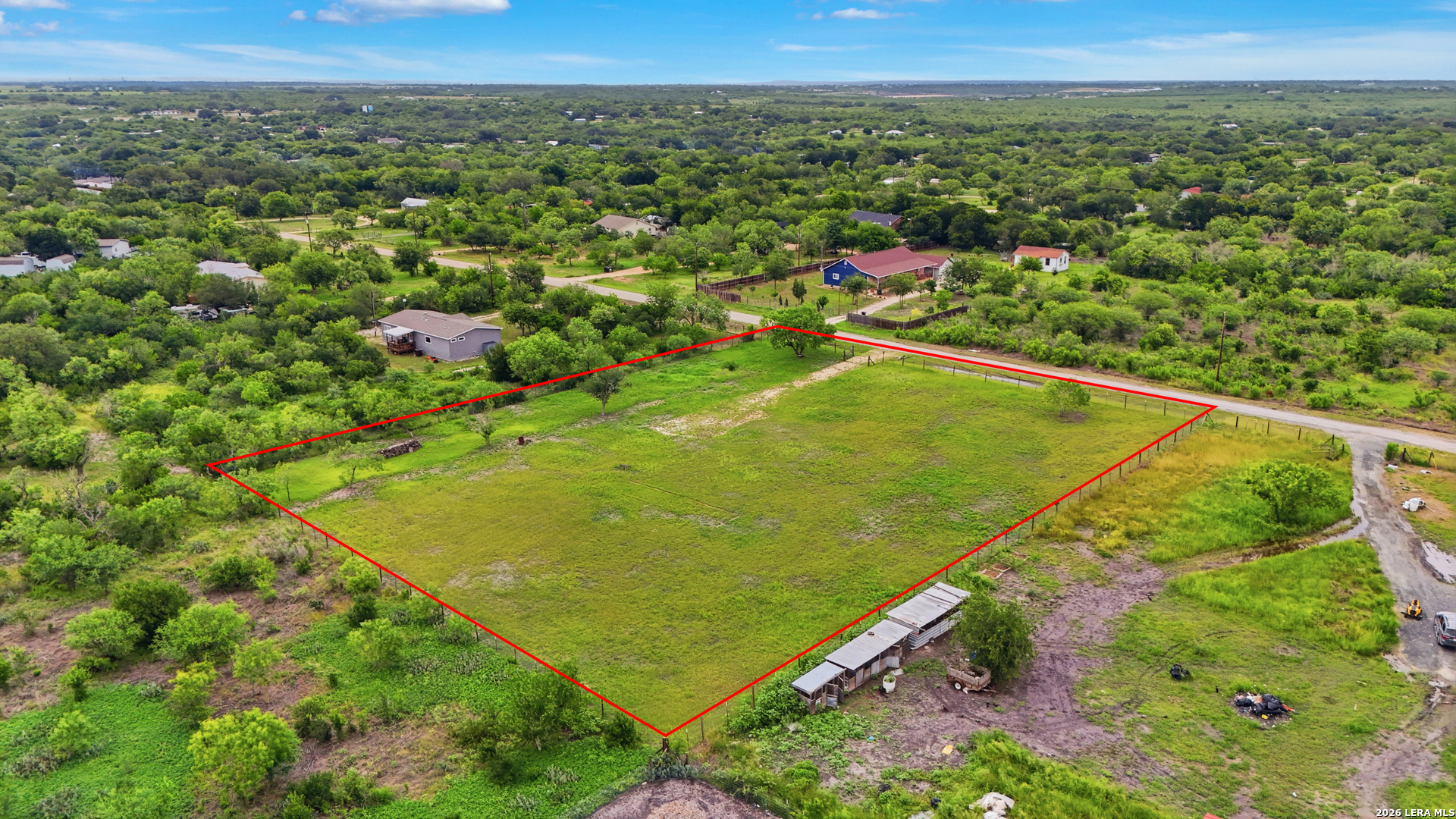 Aerial view of a grassy fenced parcel outlined in red, with outbuildings and nearby homes.