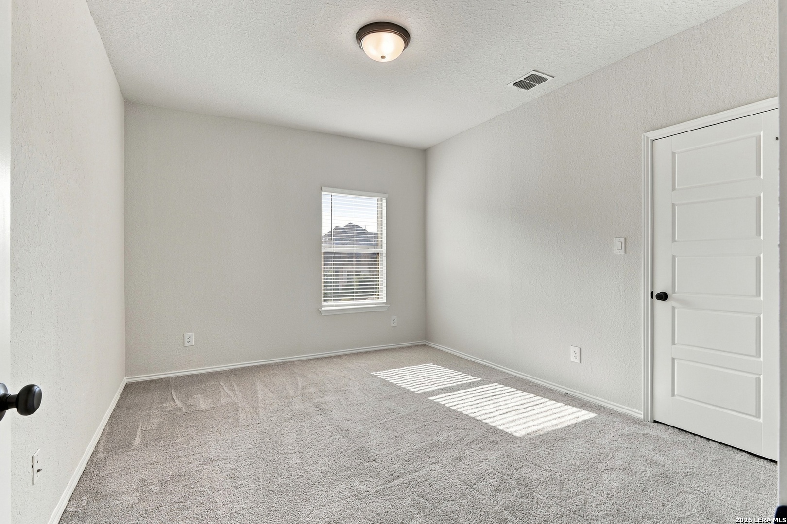 Empty bedroom with gray carpet, a window with blinds, and sunlight across the floor.