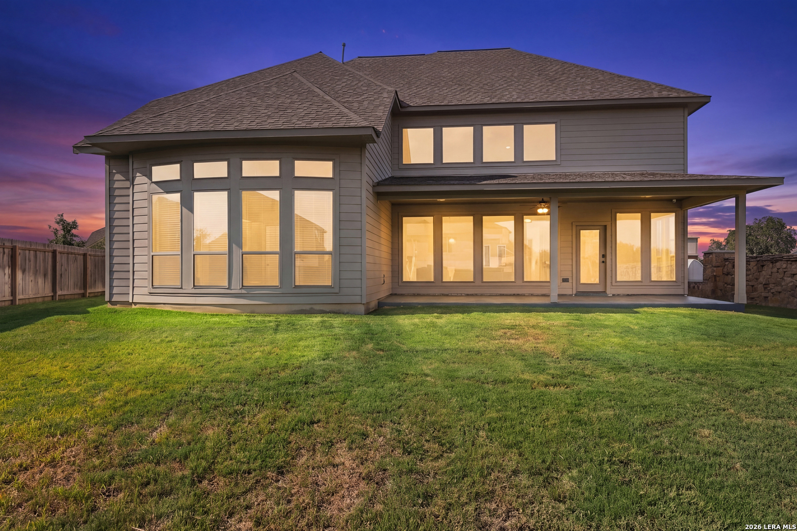 Two-story gray home with large windows, a covered patio, and broad lawn at sunset.