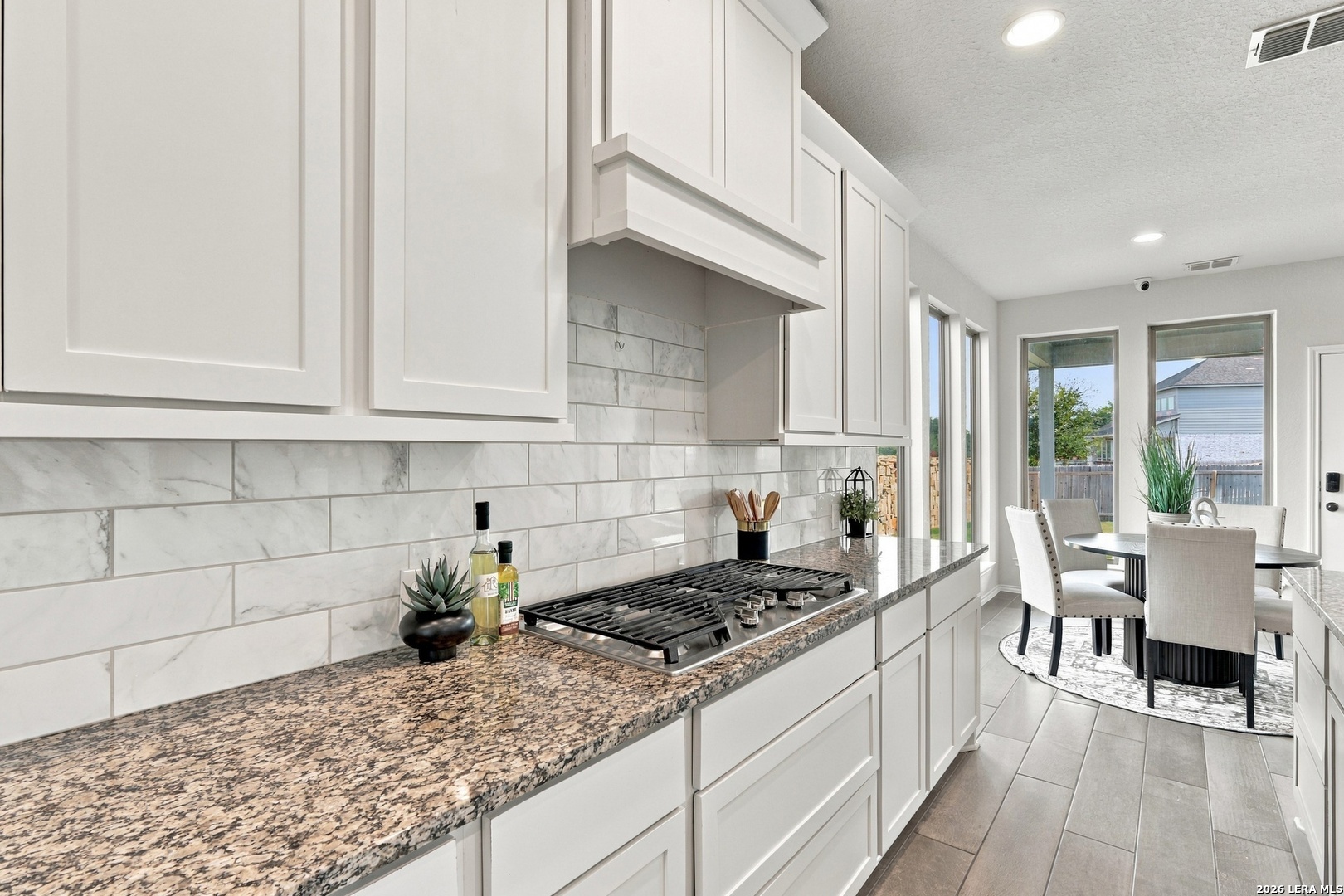 Bright kitchen with white cabinets, granite counters, a large gas cooktop, and a dining area by tall windows.
