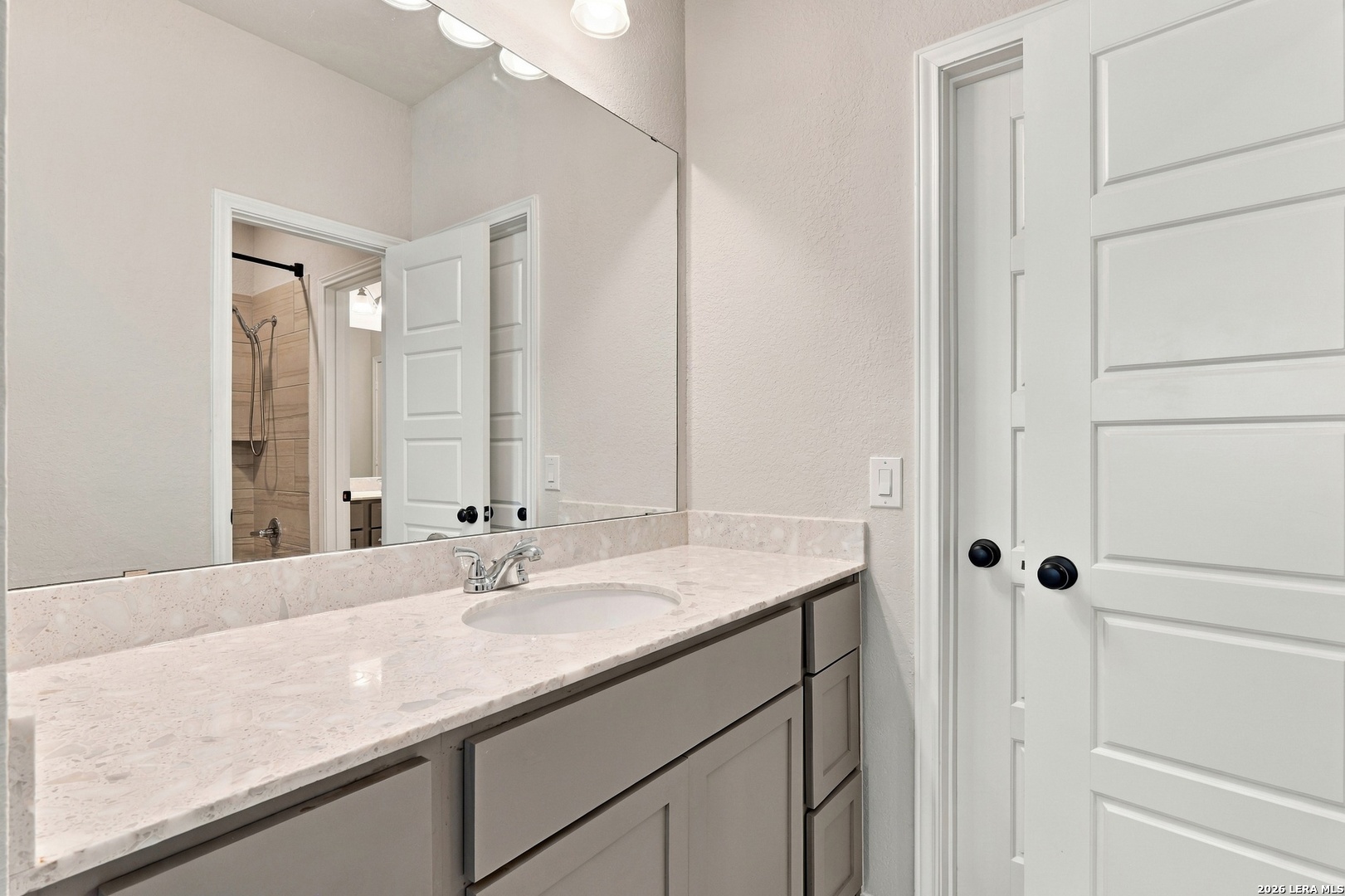 Bathroom vanity with pale stone countertop and large mirror; tiled shower visible through the doorway.