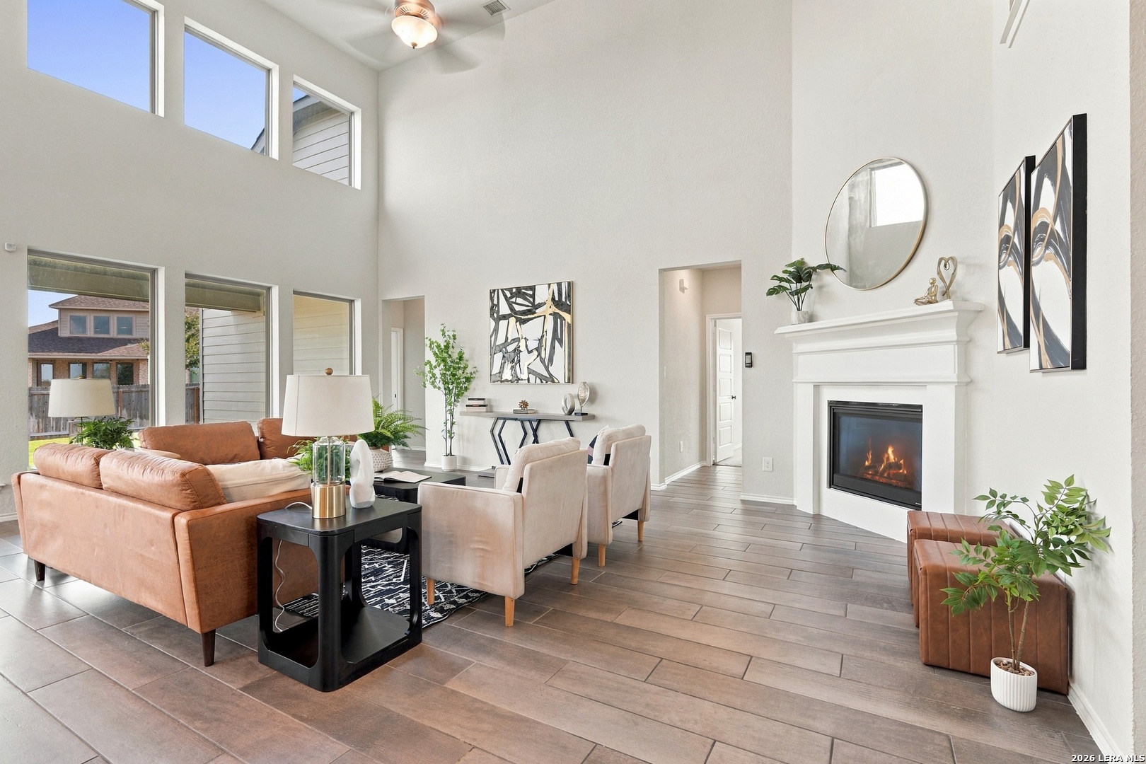 Staged living room with a double-height ceiling, tall windows, tan seating, and a white fireplace.
