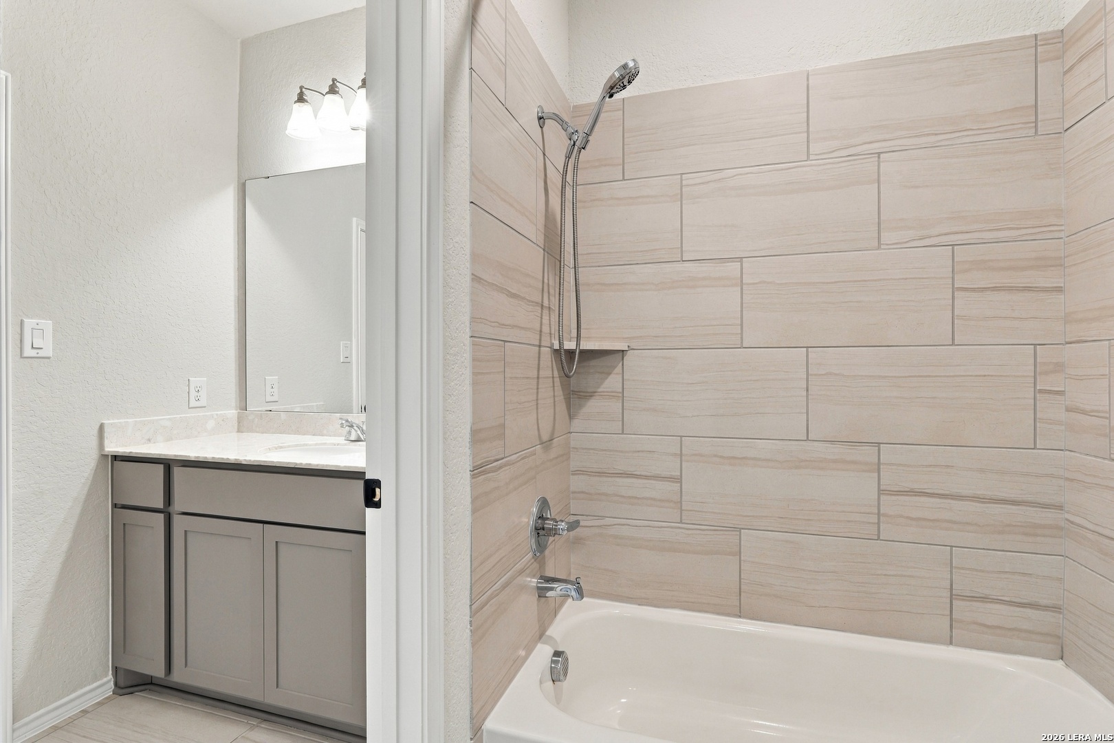 Bathroom with beige tiled tub surround, handheld showerhead, and gray vanity.