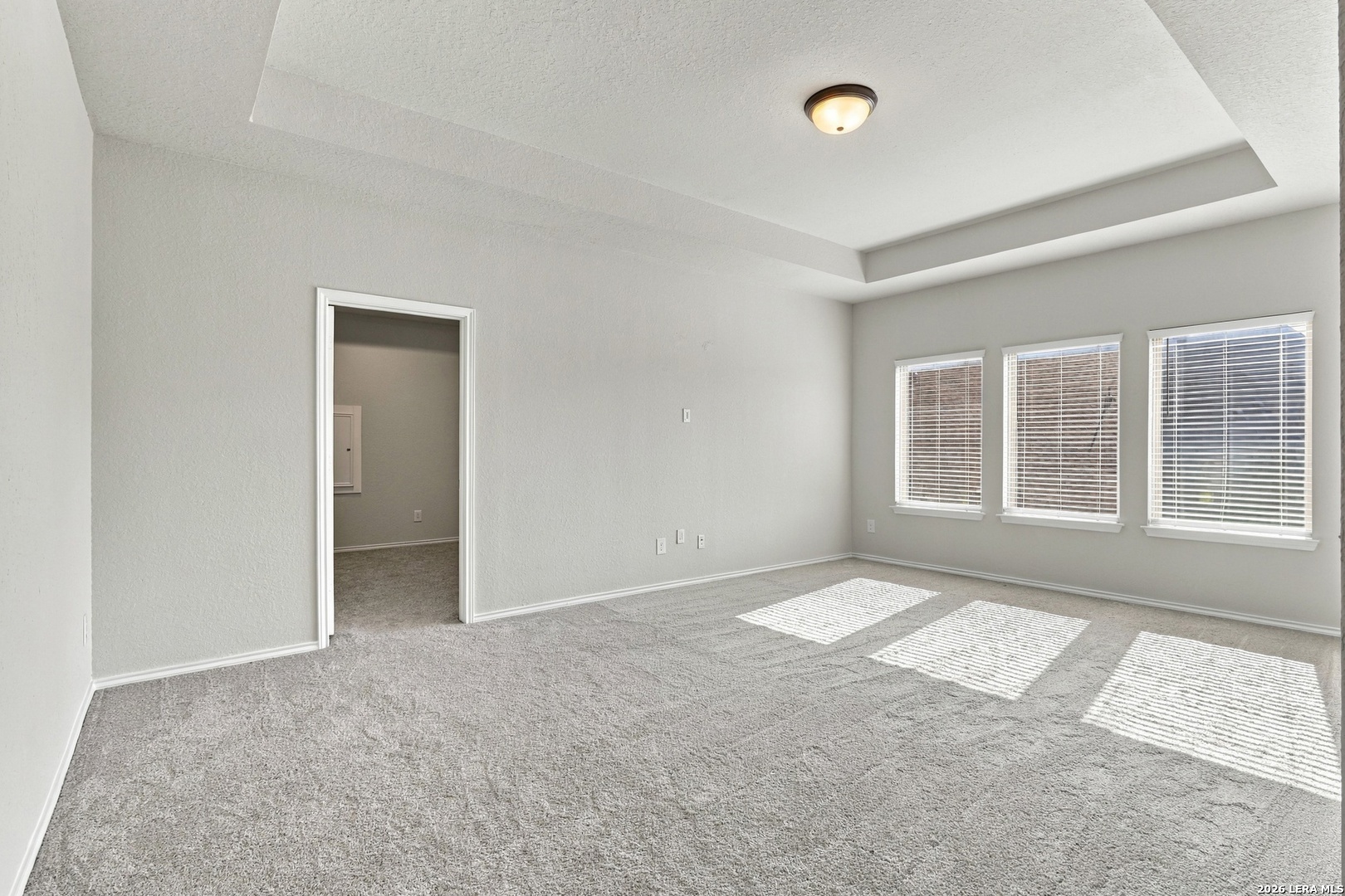 Empty carpeted room with a tray ceiling and three sunlit windows with blinds.