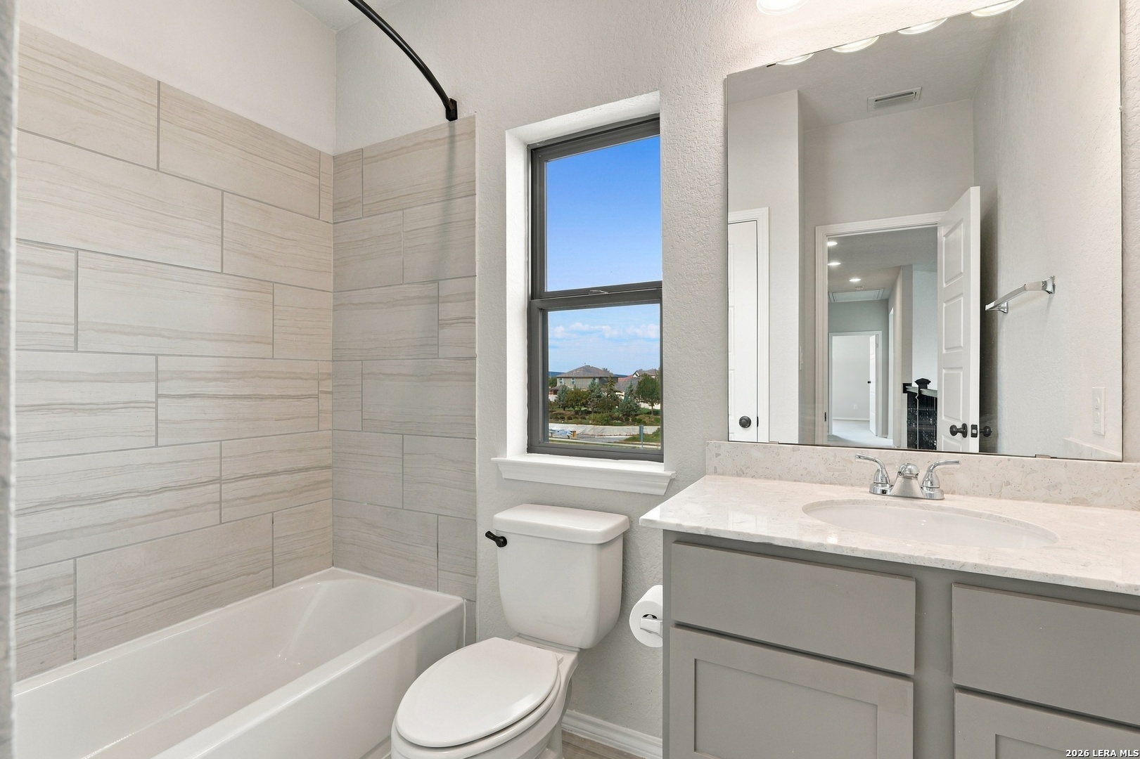 Bathroom with a tiled bathtub surround, white vanity, and window overlooking neighboring homes.