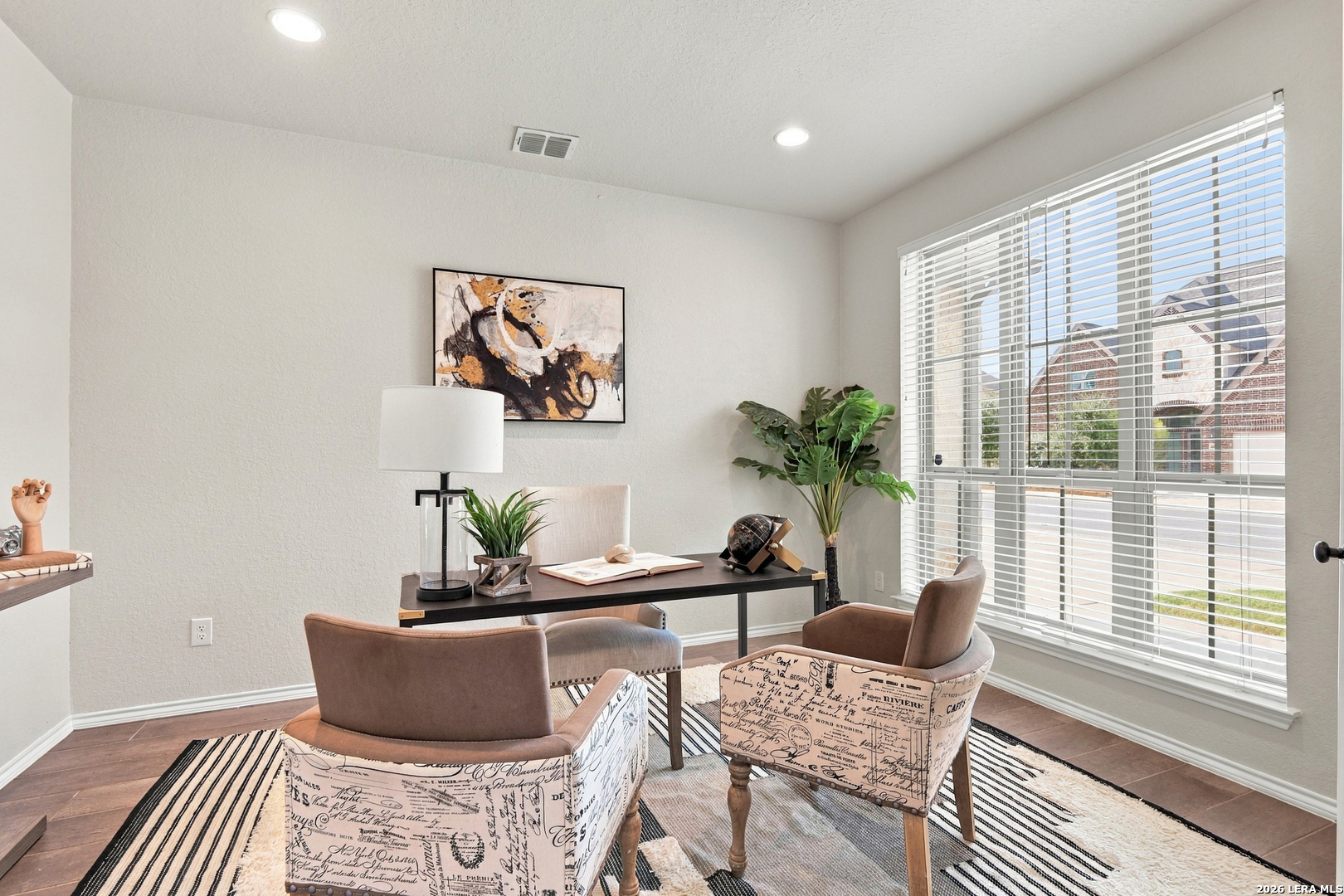 Staged home office with a desk, upholstered chairs, and a large window with white blinds.