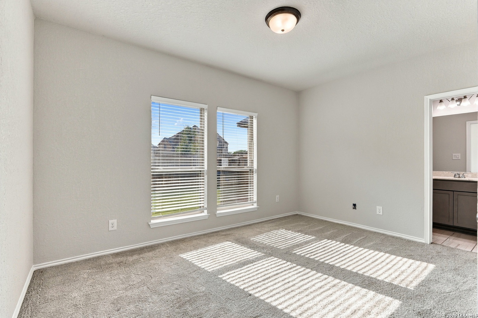 Vacant carpeted bedroom with two windows and sunlight striping the floor; bathroom vanity visible through doorway.