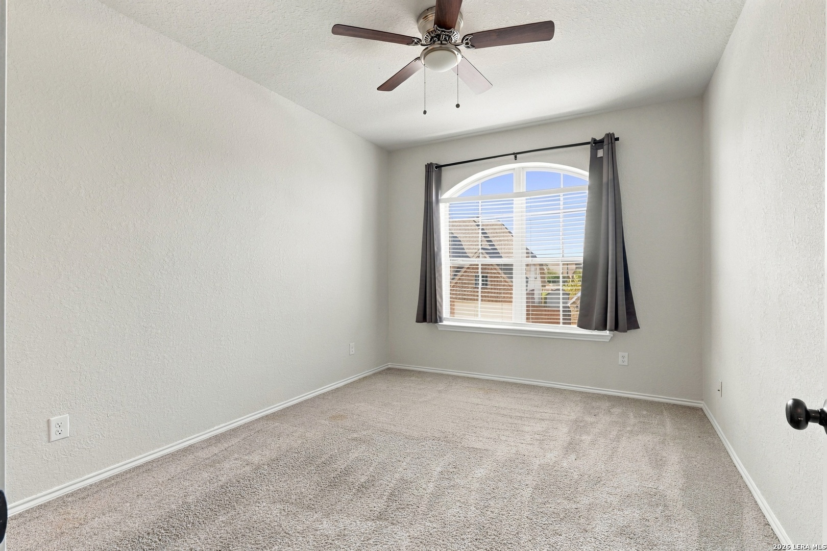 Empty carpeted bedroom with a ceiling fan and arched window with blinds and gray curtains.