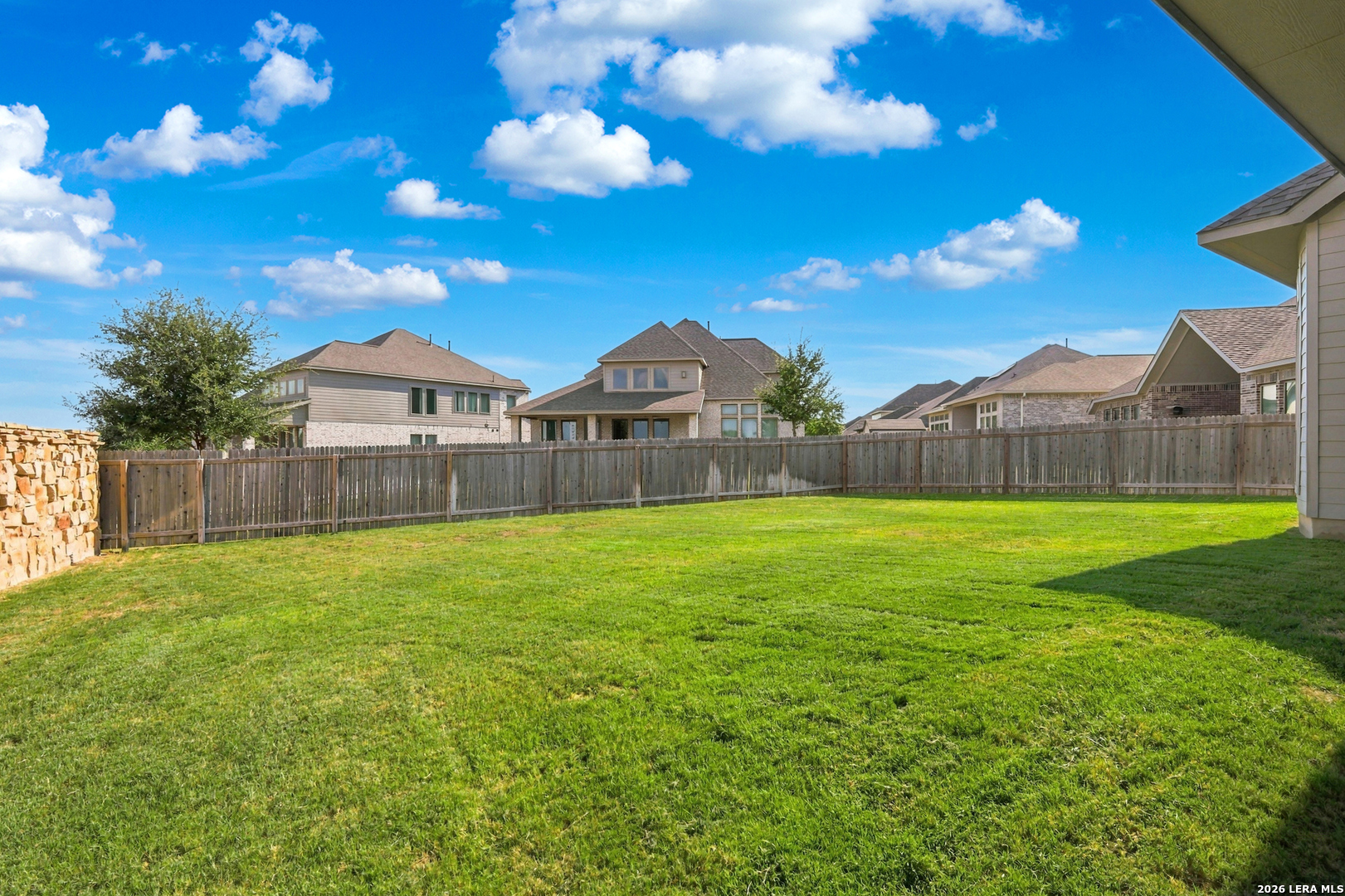 Fenced backyard with a broad green lawn, stone wall, and neighboring homes beyond the fence.