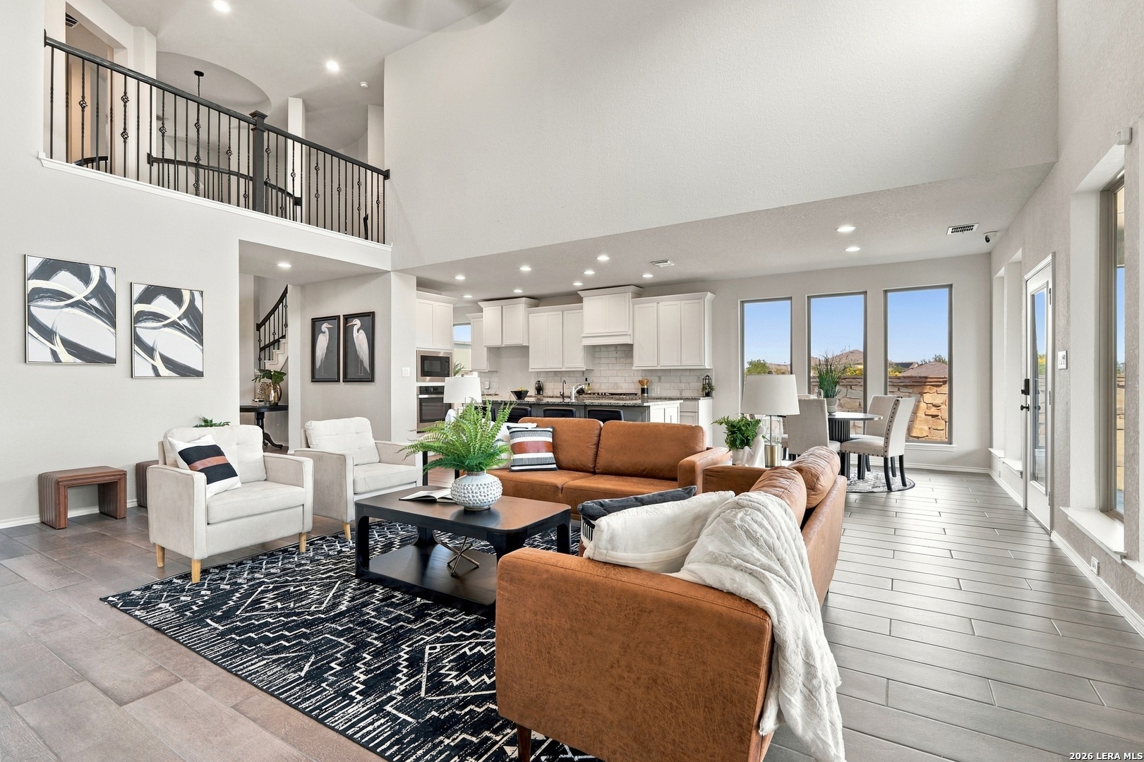Staged living room with a brown sectional, tall ceiling, iron balcony railing, and open kitchen.