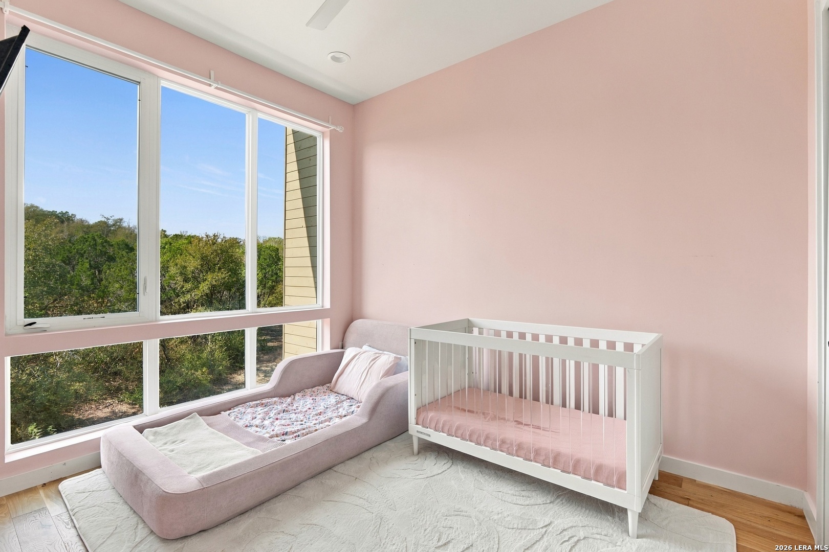 Pink nursery with a white crib and blush toddler bed beside large windows overlooking trees.