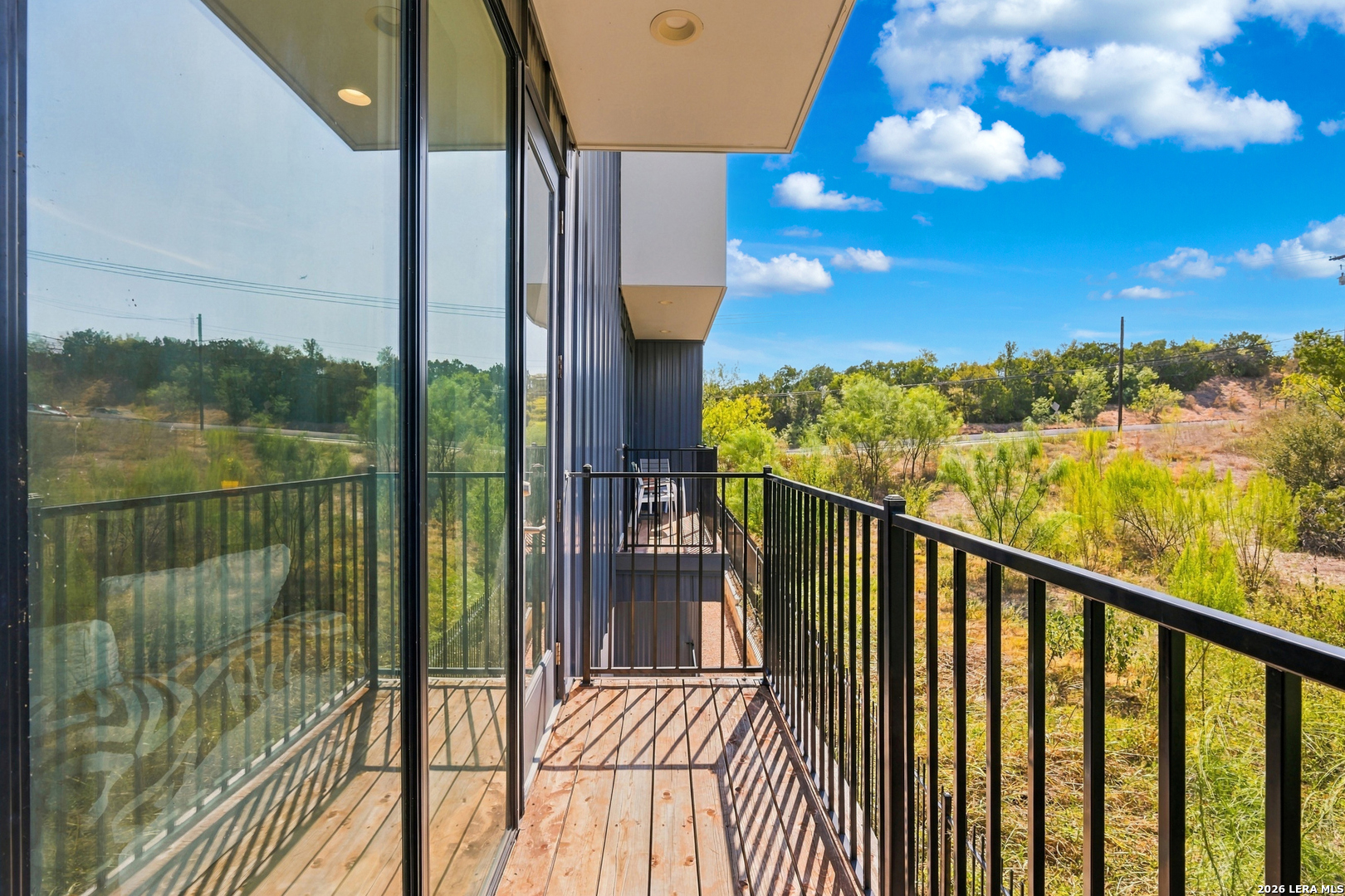 Wooden balcony with black railings beside floor-to-ceiling glass doors, overlooking trees and brush.