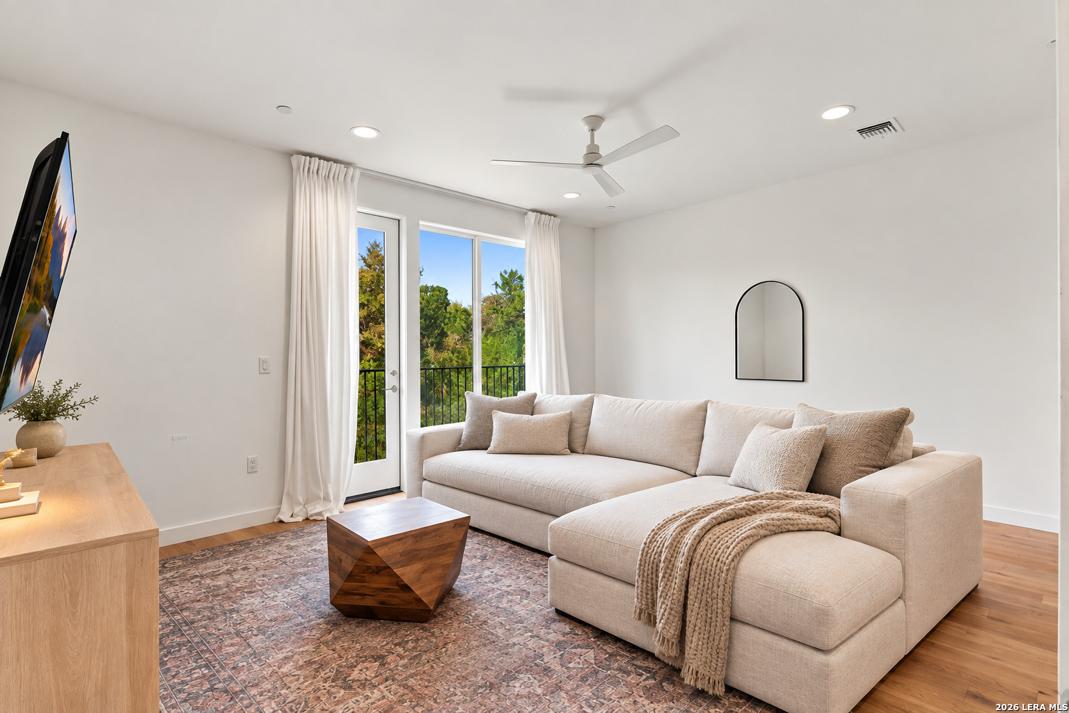 Living room with a cream sectional, patterned rug, wood coffee table, and sliding door to a tree-lined balcony.