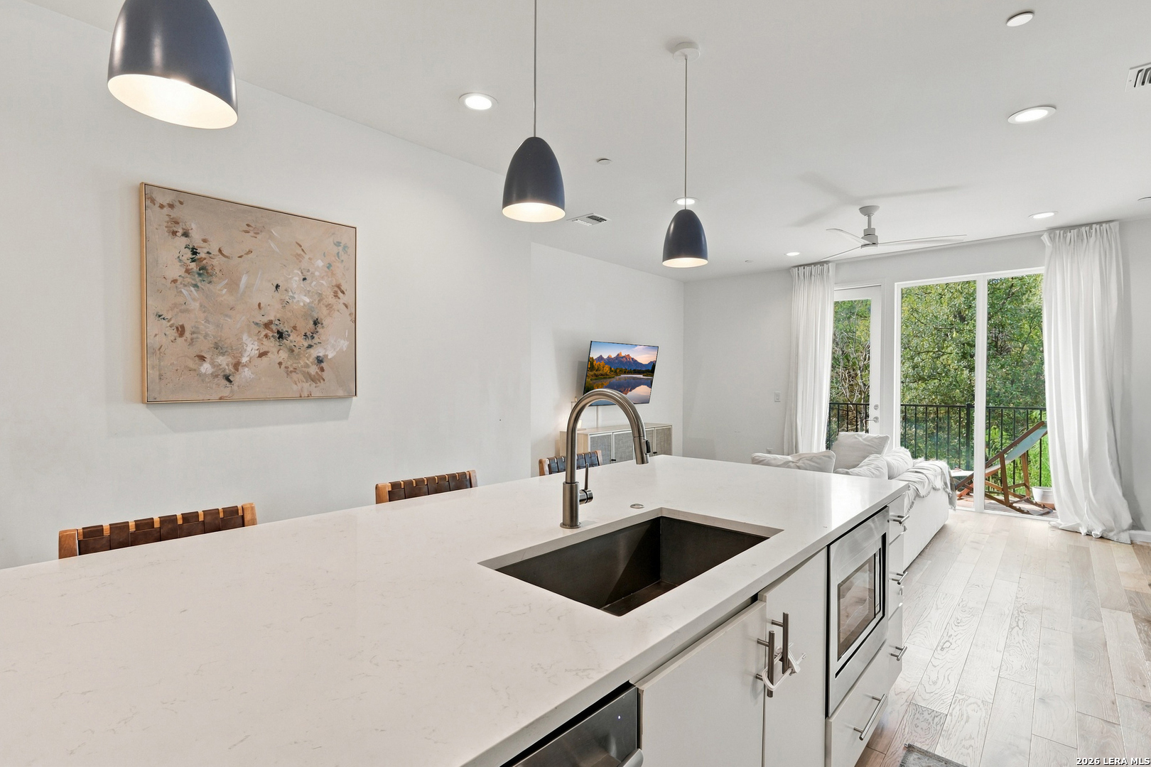 Open-plan kitchen with a large white island and black sink, opening onto a leafy balcony.